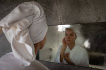 Sobre la vista del hombro de una joven mujer caucásica usando un albornoz con su cabello envuelto en una toalla, mirando en el espejo en un baño moderno . - foto de stock