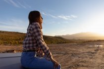 Крупним планом вид з боку молодої жінки, що сидить на капоті пікапа, насолоджуючись видом на заході сонця під час зупинки в дорожній поїздці — стокове фото