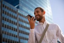 Vista frontal de cerca de un joven caucásico sonriente hablando en un teléfono inteligente que lo sostiene frente a su cara en una calle de la ciudad. Nómada digital en movimiento . - foto de stock