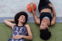 Повышен крупный план двух молодых взрослых смешанных гоночных сестер в спортивной одежде, лежащих на земле, смотрящих друг на друга и улыбающихся, одна держит баскетбольный мяч — стоковое фото
