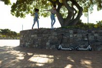 Vista lateral de dos jóvenes hermanas de raza mixta adultas sonrientes caminando a lo largo de una pared en un parque urbano, con sus scooters eléctricos estacionados debajo de ellas - foto de stock