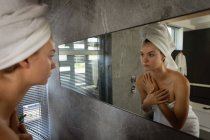 Vue de côté gros plan d'une jeune femme caucasienne portant une serviette de bain et avec ses cheveux enveloppés dans une serviette, regardant dans le miroir dans une salle de bain moderne . — Photo de stock