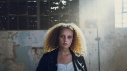 Vista frontal de uma jovem caucasiana com cabelo encaracolado usando jaqueta de couro enquanto olha atentamente para a câmera com luz solar sobre ela dentro de um armazém vazio — Fotografia de Stock