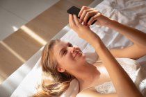 Крупним планом посміхаючись молода кавказька блондинка лежачи на спині в ліжку за допомогою смартфона. — стокове фото