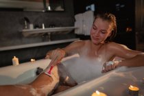 Vorderansicht einer lächelnden jungen kaukasischen Frau, die in der Badewanne sitzt und sich die Beine rasiert, mit brennenden Kerzen rund um die Badewanne. — Stockfoto