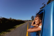 Side view of a young mixed race woman leaning out of the front passenger side window of a pick-up truck smiling, as it drives down the highway on a road trip — Stock Photo