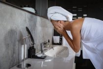 Side view of a young Caucasian woman washing her face, wearing a bath towel and with her hair wrapped in a towel, in a modern bathroom. — Stock Photo
