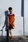 Vue de face d'une jeune femme métissée portant une robe, appuyée contre un mur dans une rue urbaine au soleil tenant une planche à roulettes — Photo de stock