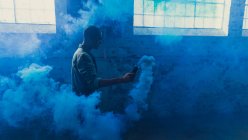 Seitenansicht eines jungen hispanisch-amerikanischen Mannes, der eine graue Jacke über einem weißen Hemd trägt und eine Rauchmaschine in der Hand hält, die blauen Rauch in einer leeren Lagerhalle produziert — Stockfoto