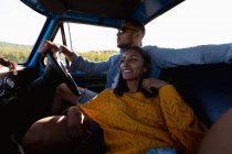 Vista lateral de cerca de una joven pareja mixta sentada en su camioneta durante un viaje por carretera. El hombre está conduciendo y la mujer se apoya en él y sonríe - foto de stock