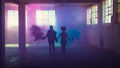 Vista frontal de la silueta de una pareja con fabricantes de humo que producen humo violeta y azul mientras se toman de la mano y corren dentro de un almacén vacío - foto de stock