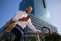 Vue de face gros plan d'un jeune homme caucasien souriant utilisant un smartphone, assis sur son vélo dans une rue de la ville. Nomade numérique en mouvement . — Photo de stock