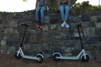 Vista frontale sezione bassa di due sorelle sedute su un muro in un parco urbano, con i loro scooter elettrici parcheggiati sotto di loro — Foto stock