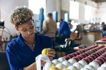 Front view close up of a young mixed race man sitting polishing cricket balls at the end of the production line at a cricket ball factory. In the background colleagues are operating machines at workbenches. — Stock Photo