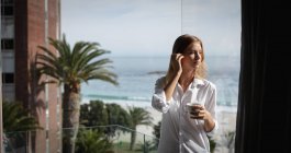 Vista frontale di una giovane donna caucasica con una camicia bianca in piedi su un balcone che regge una tazza di caffè e distoglie lo sguardo, palme e spiaggia sullo sfondo . — Foto stock