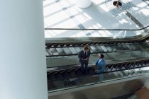 Vista de ángulo alto de hombres de negocios que interactúan entre sí mientras se mueven arriba en escaleras mecánicas en la oficina . - foto de stock