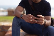 Vorderseite Mittelteil eines Mannes, der draußen an einer Wand sitzt und ein Smartphone in der Sonne benutzt — Stockfoto