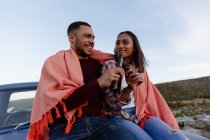 Close up front view of a young mixed race couple sitting outside in the back of their pick-up truck with a blanket over their shoulders, holding bottles of beer and making a toast during a stop off on a road trip. — Stock Photo