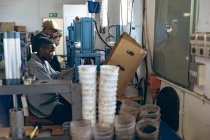 Vista lateral de una amplia gama de edad de trabajadores afroamericanos sentados y operando máquinas en una fábrica de bolas de cricket, rodeado de equipos y materiales . - foto de stock