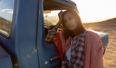 Portrait of a thoughtful young mixed race woman with a blanket over her shoulders, leaning on the door of a pick-up truck looking straight to camera at sundown, during a stop off on a road trip — Stock Photo