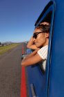 Vista lateral cercana de una joven mujer de raza mixta que lleva gafas de sol asomadas por la ventana del lado delantero del pasajero de una camioneta sonriente, mientras conduce por la carretera en un viaje por carretera - foto de stock