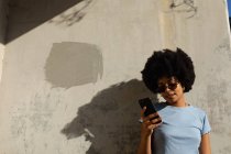 Front view close up of a young mixed race woman wearing sunglasses standing against a wall using her smartphone in the sun — Stock Photo