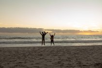 Vista trasera de un hombre y una mujer caucásicos maduros saltando juntos con sus brazos en el aire en una playa, frente al mar al atardecer - foto de stock