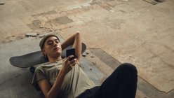 High angle view of a young Hispanic-American man with piercings wearing a dark grey shirt and beanie lying on the floor with head on a skateboard and using a mobile phone — Stock Photo