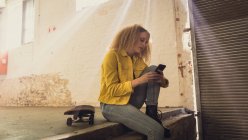 Вид збоку молода кавказька жінка з Кучеряве волосся носити жовту куртку над сірої сорочці сидить поруч з скейтбордом при використанні мобільного телефону всередині порожній склад — стокове фото