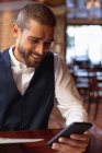 Vista frontal de cerca de un joven caucásico sonriente usando su teléfono inteligente sentado en una mesa dentro de un café. Nómada digital en movimiento
. - foto de stock