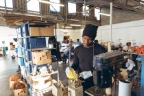 Frontansicht eines jungen afrikanisch-amerikanischen Mannes mit Hut, der an einer Werkbank steht und in einer Werkstatt in einer Cricketfabrik Gummi in eine Form gießt, im Hintergrund arbeiten Kollegen an anderen Teilen der Produktionslinie. — Stockfoto