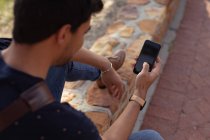 Над плечовим видом молодого змішаного чоловіка, який сидить на вулиці на стіні, використовуючи смартфон на сонці — стокове фото