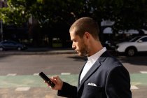 Vista lateral de perto de um jovem caucasiano vestindo um casaco usando seu smartphone em pé em uma rua da cidade. Digital Nomad em movimento . — Fotografia de Stock