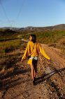 Вид спереду на молоду змішану жінку, яка насолоджується прогулянкою по стежці через сонячний сільський пейзаж до гір на горизонті. Вона носить шорти, з жовтим верхом з сумочкою і камерою . — стокове фото