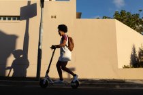 Seitenansicht einer jungen Frau mit gemischtem Rennen, die einen Rucksack trägt und auf einem Motorroller in einer städtischen Straße in der Sonne fährt — Stockfoto
