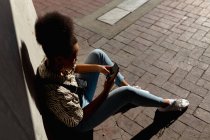 Высокий крупный план молодой смешанной расы женщины, сидящей на тротуаре с помощью смартфона на солнечной городской улице — стоковое фото