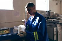 Vue latérale rapprochée d'un jeune homme de race mixte portant des gants et des salopettes préparant un mélange dans une usine fabriquant des balles de cricket, avec un équipement visible en arrière-plan . — Photo de stock