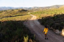 Вид спереду молодої змішаної раси жінки, що йде по стежці через сонячний сільський пейзаж до гір на горизонті. Вона носить шорти, з жовтим верхом і сумочкою . — стокове фото