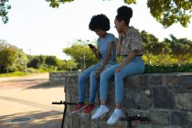 Вид збоку на двох усміхнених молодих сестер змішаної раси, які сидять на стіні в міському парку, використовуючи смартфон та обмінюючись навушниками для прослуховування музики, з їх електричними скутерами, припаркованими під ними — стокове фото