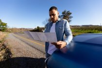 Vista frontal de cerca de un joven hombre de raza mixta apoyado en la capucha de una camioneta que lee un mapa durante una parada al borde de la carretera en un viaje por carretera . - foto de stock