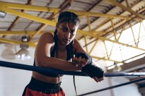 Низкий угол обзора женского боксера, опирающегося на веревки и смотрящего на камеру в боксерском ринге. Сильная женщина-боец в боксёрском зале тяжело тренируется . — стоковое фото