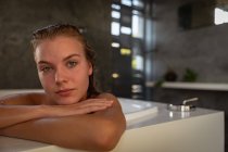 Retrato close-up de uma jovem mulher caucasiana sentada na banheira e olhando diretamente para a câmera em um banheiro moderno . — Fotografia de Stock