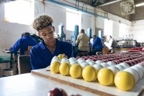 Front view of a young mixed race man sitting polishing cricket balls at the end of the production line at a cricket ball factory. In the background colleagues are operating machines at workbenches. — Stock Photo