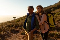 Side view close up of a mature Caucasian man and woman wearing backpacks stopping to enjoy the scenery together at sunset during a hike. — Stock Photo