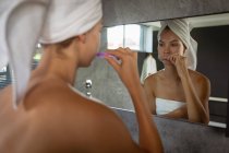 Über die Schulter einer jungen kaukasischen Frau, die sich die Zähne putzt, ein Badetuch trägt und ihr Haar in ein Handtuch gehüllt hat, das sich im Spiegel eines modernen Badezimmers spiegelt. — Stockfoto