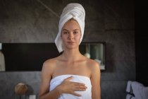 Retrato de uma jovem caucasiana vestindo uma toalha de banho e com o cabelo envolto em uma toalha, olhando diretamente para a câmera em um banheiro moderno . — Fotografia de Stock