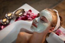Vista laterale ravvicinata di una giovane donna caucasica sdraiata in un bagno con maschera facciale, con petali e candele accese intorno alla vasca da bagno . — Foto stock