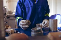 Front view mid section of man wearing gloves and overalls holding the core of a ball and checking it at a factory making cricket balls, surrounded by equipment. — Stock Photo