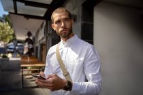 Front view close up of a young Caucasian man using a smartphone, standing on the pavement in a city street and looking away. Digital Nomad on the go. — Stock Photo