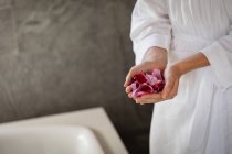 Средняя часть женщины в халате с горсткой лепестков роз, стоящей рядом с ванной в современной ванной комнате . — стоковое фото